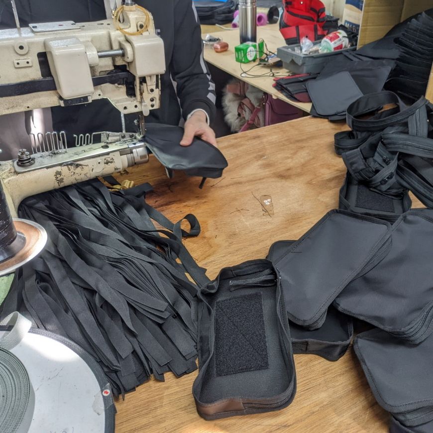 Industrial sewing workstation with technician assembling black fabric components, including Velcro-equipped pieces, illustrating tactical gear production prior to quality inspection